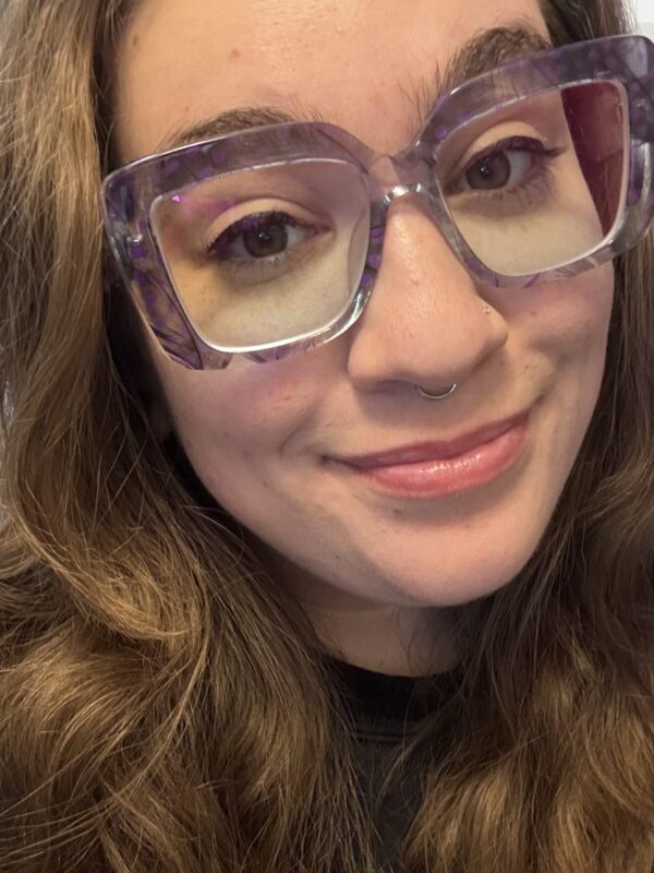Selfie of a smiling person with long, wavy brown hair, oversized clear purple glasses, and a septum ring, wearing a black shirt.